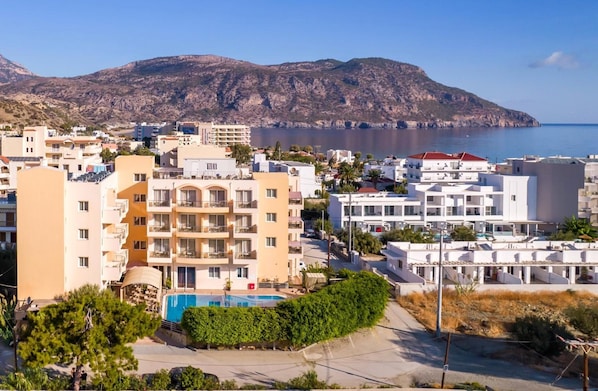 A coastal residential area with multi-story buildings, a swimming pool, and a mountainous backdrop. - Nereides (Karpathos)