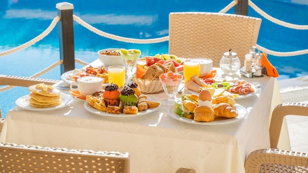 Ein Frühstücksbuffet mit verschiedenen Speisen, im Hintergrund ein Schwimmbecken und Korbstühle um den Tisch.