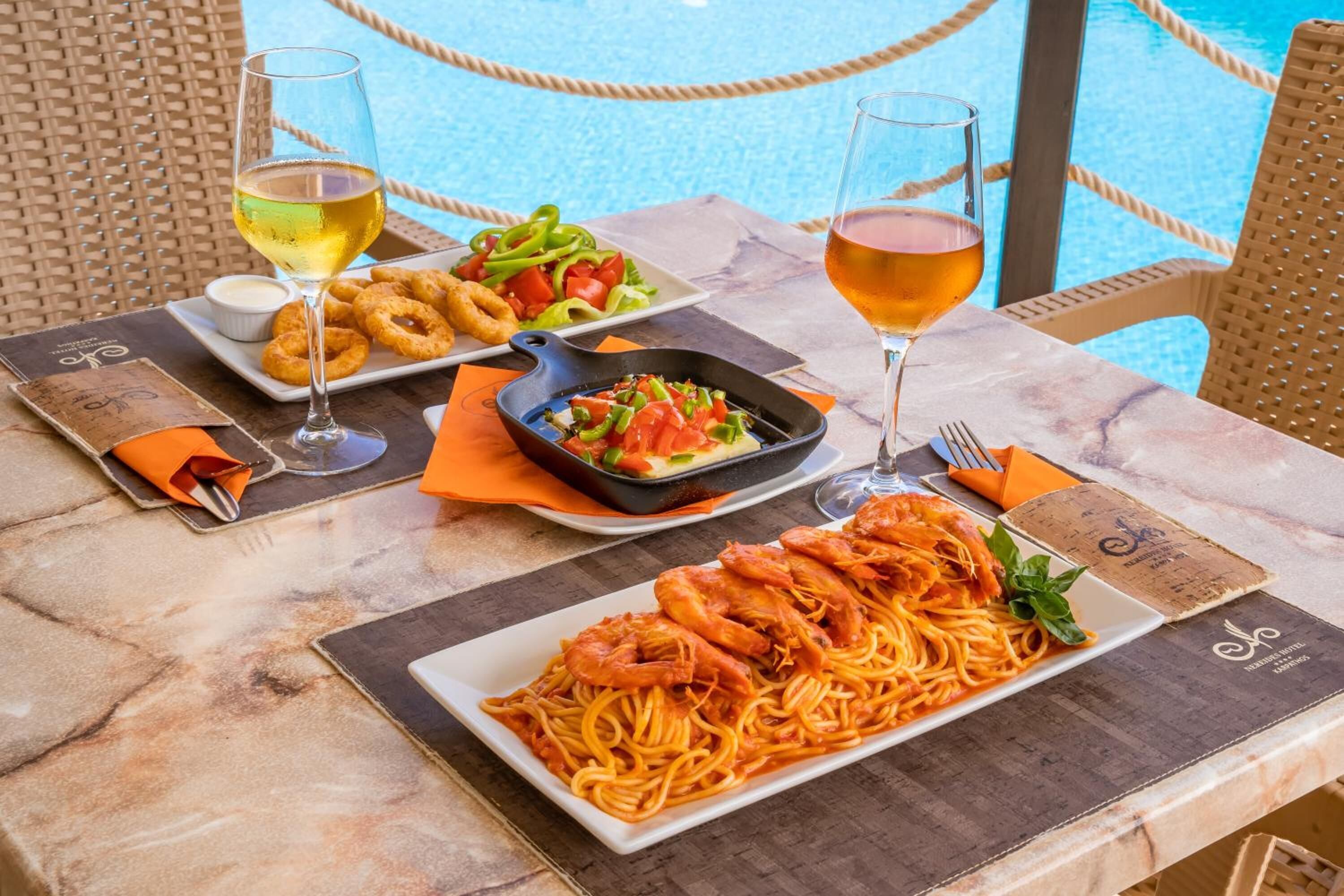 A dining table with seafood pasta, a salad, and fried calamari, accompanied by two glasses of wine, set against a poolside view.