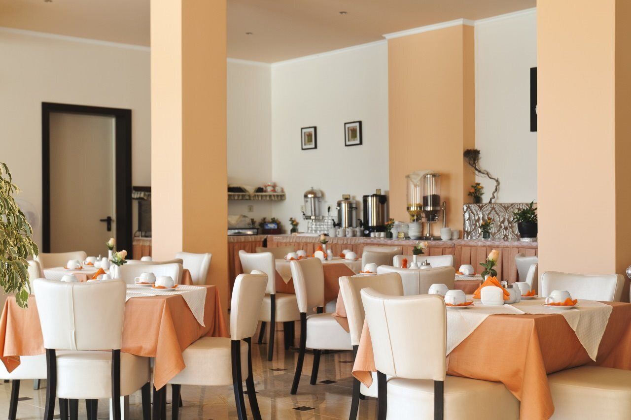 A restaurant with white chairs and tables, orange tablecloths, and a bar area with kitchen equipment.