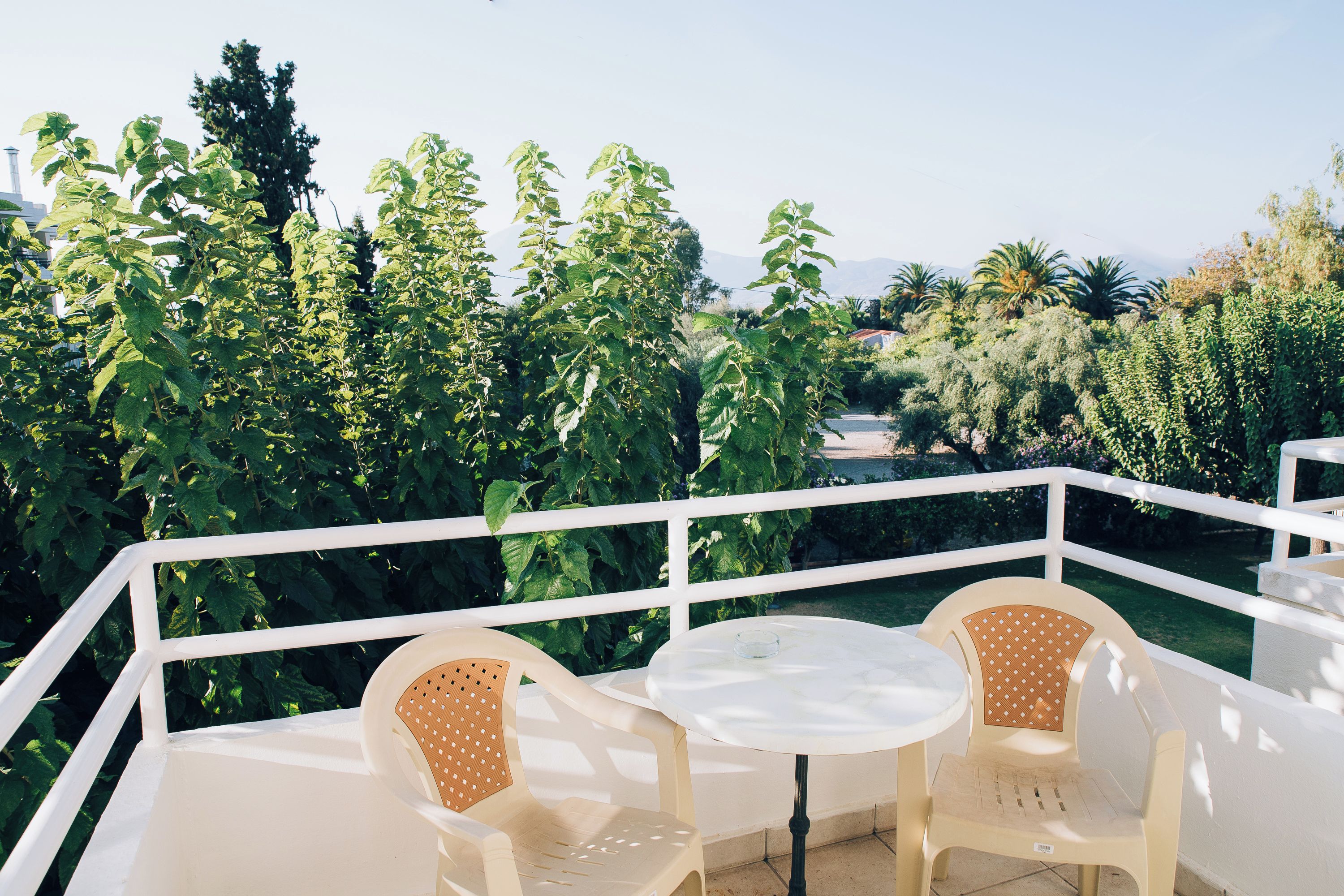 An outdoor seating area with white chairs and a table, surrounded by lush greenery and a clear sky.