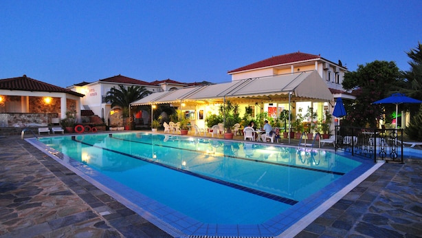 A swimming pool with lounge chairs and umbrellas, surrounded by stone flooring and a building with a red roof. - Hotel Metaxa (Zakynthos)