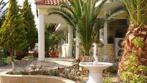 A garden with a fountain, palm trees, and a building with a tiled roof. - Hotel Metaxa (Zakynthos)
