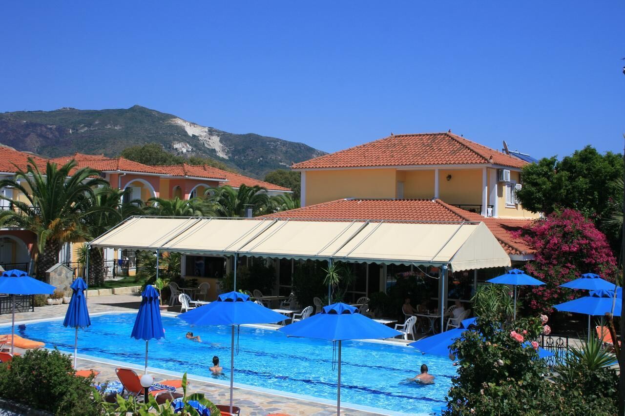 A pool area with blue umbrellas and lounge chairs, surrounded by buildings with red-tiled roofs and a mountain in the background.
