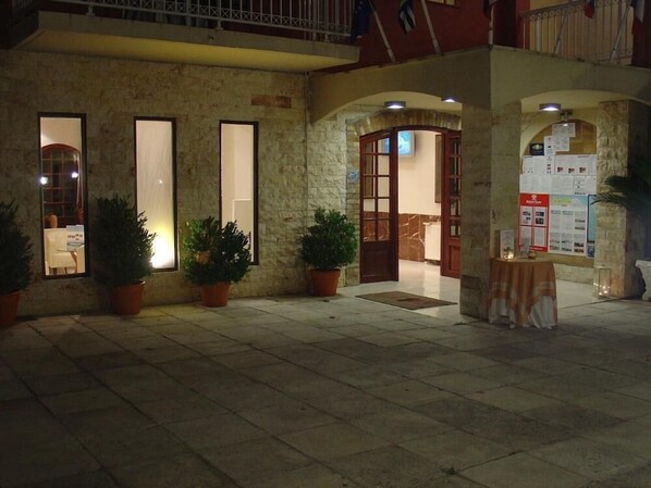 A building with stone walls, large windows, and a door leading inside. There are potted plants outside and a table with a sign on the right side. - Oasis (Corfu)