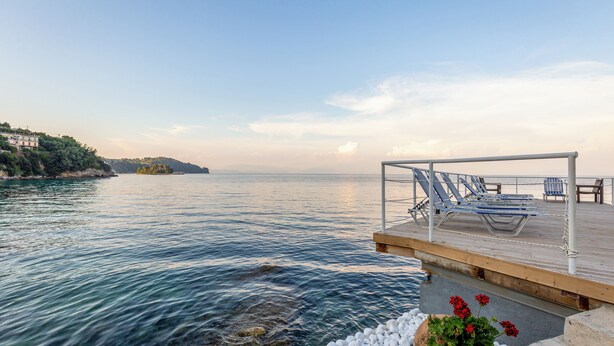 A wooden deck with lounge chairs overlooking a calm sea, a rocky shoreline, and a distant hill. - Oasis (Corfu)