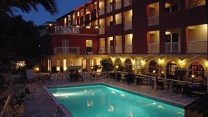 A hotel with a swimming pool, outdoor seating, and a red building facade. - Oasis (Corfu)