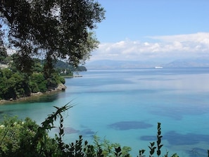 A coastal landscape with a clear view of the sea, a tree-lined shore, and a distant ship. - Oasis (Corfu)