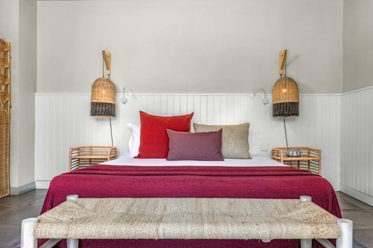 A bed with a red bedspread, two bedside lamps, and a woven bench in front.