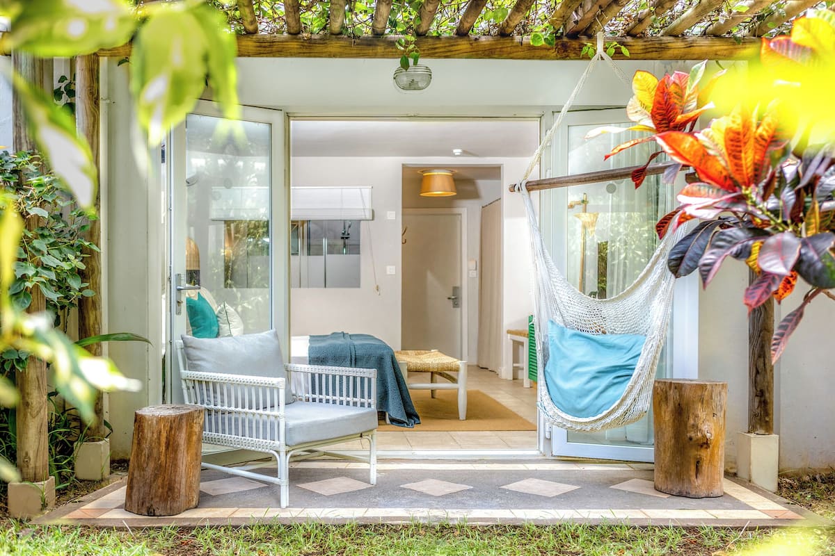 A hammock chair, a white sofa, and a wooden table in a garden area with a glass door leading to a room.