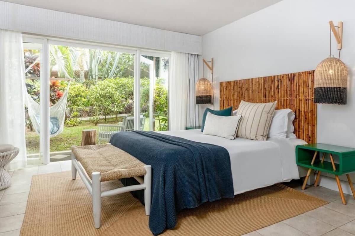 A bedroom with a bed, a bench, a green side table, and a view of a garden through large sliding doors.