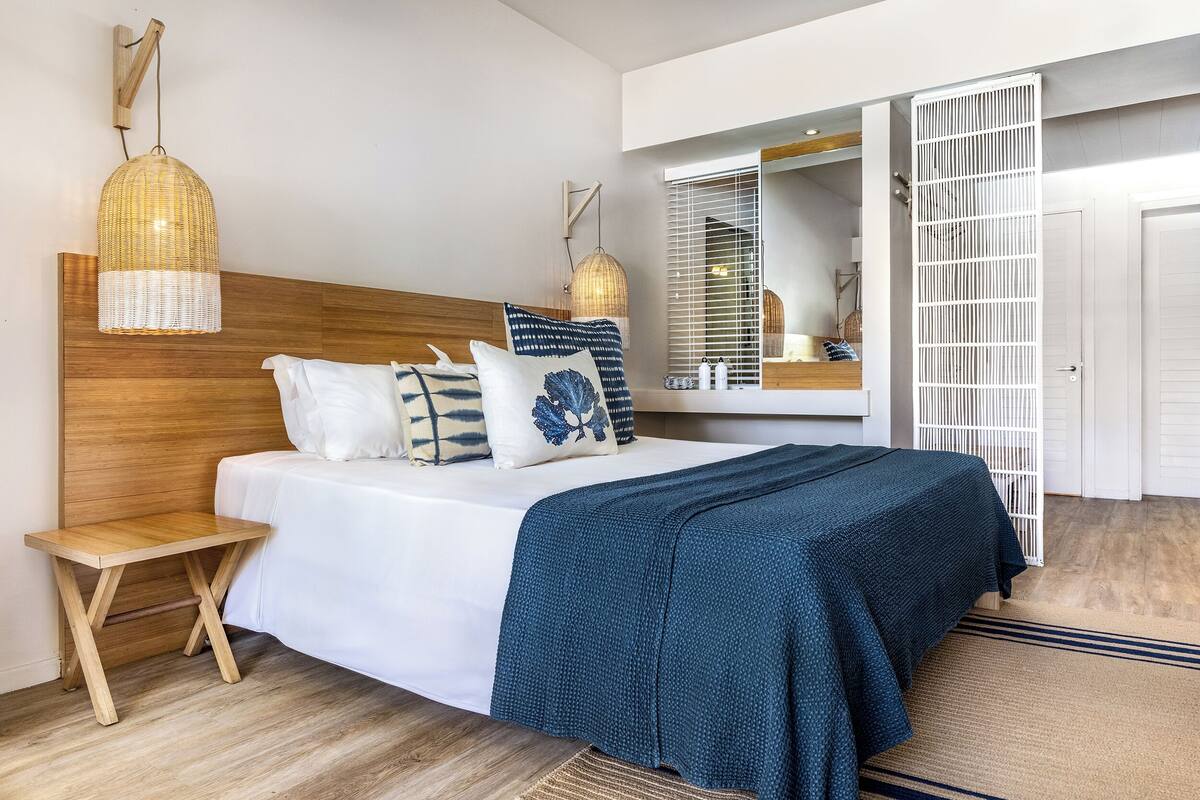 A bedroom with a wooden headboard, a bed with white bedding and a blue blanket, a small wooden nightstand, a hanging light fixture, and a large mirror with a white partition.
