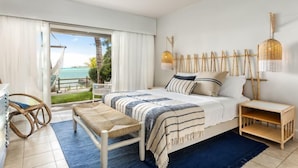A bedroom with a bed, a bench, a bedside table, a hanging light fixture, and a view of the ocean.