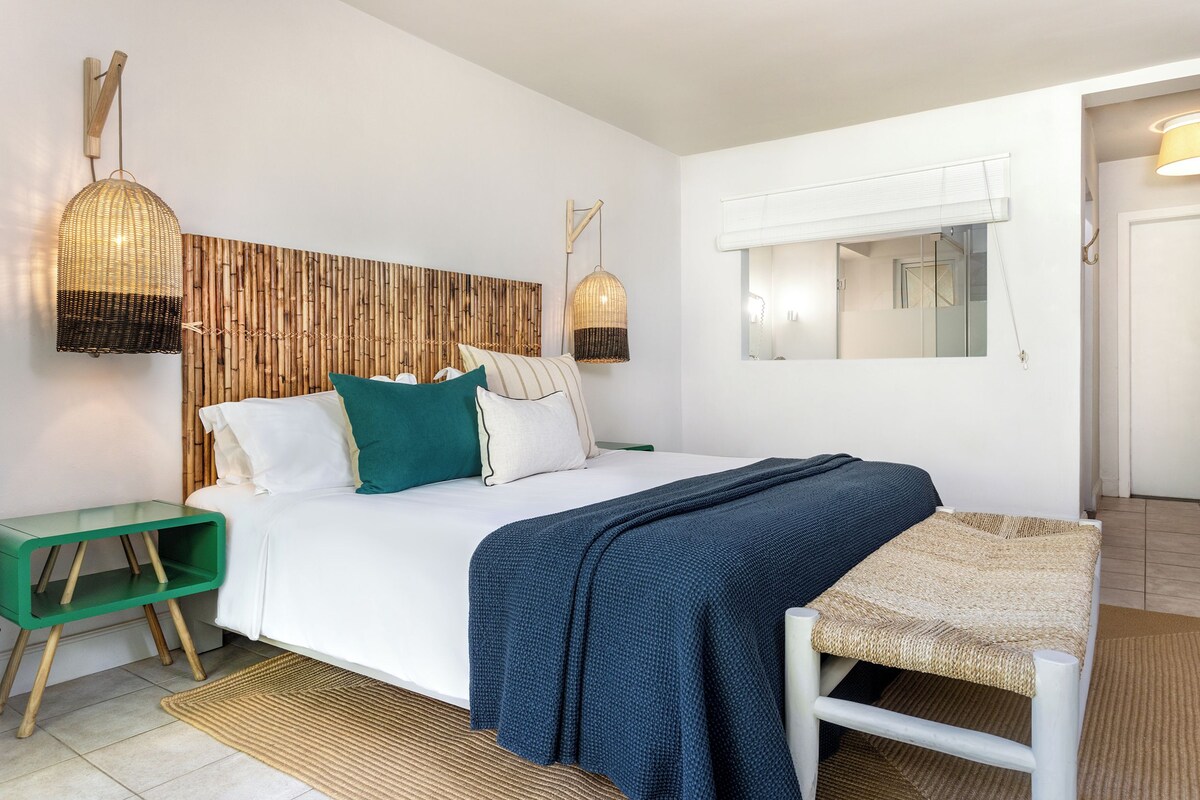 A modern bedroom with a bed, bedside tables, a woven bench, and wall-mounted lighting.