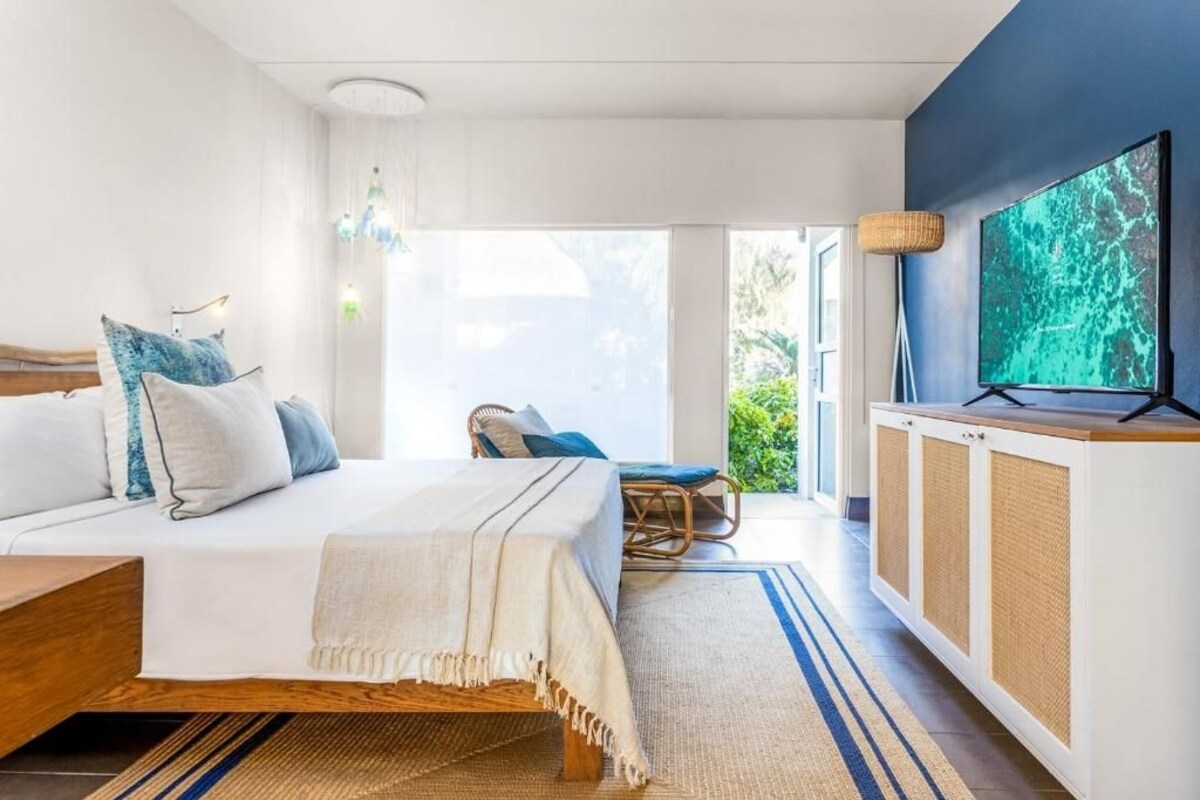 A modern bedroom with a bed, a TV, a wicker chair, and a view of greenery.