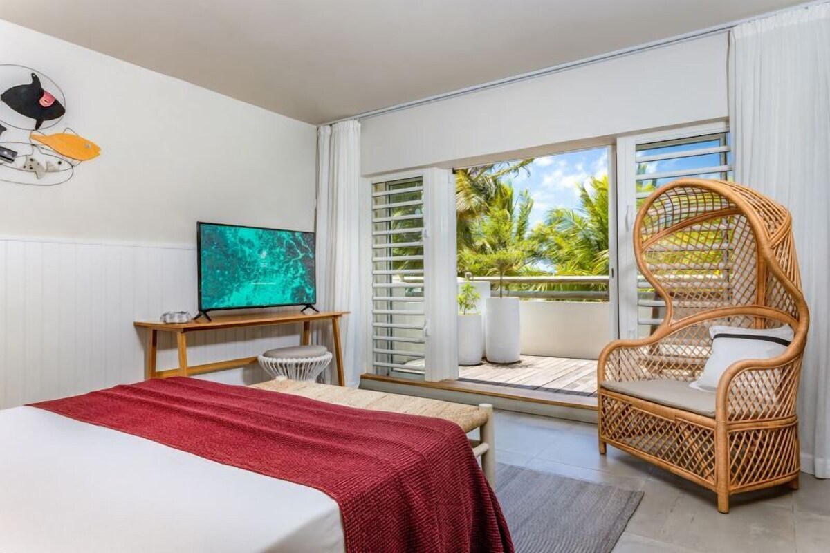 A hotel room with a bed, a television, a wicker chair, a balcony with a view of palm trees, and a wall-mounted artwork.