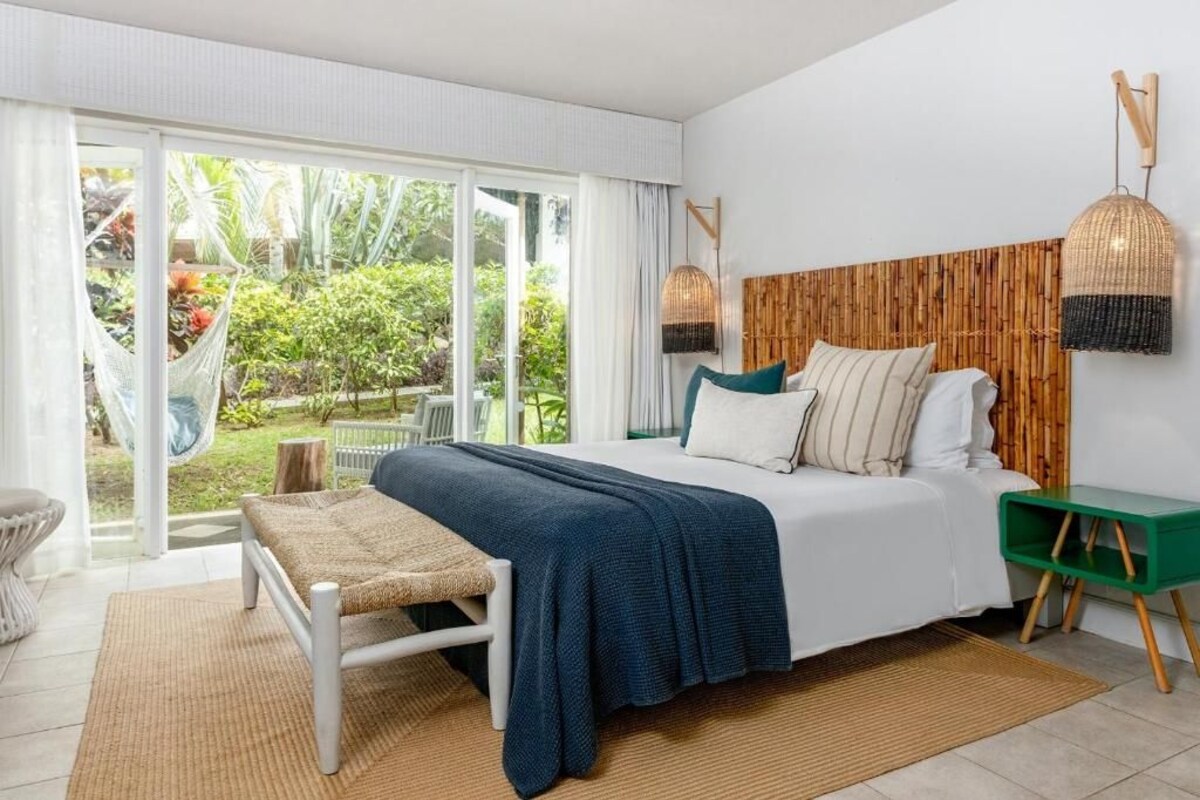 A bedroom with a large bed, a green nightstand, a woven bench, and a view of a garden through a sliding glass door.