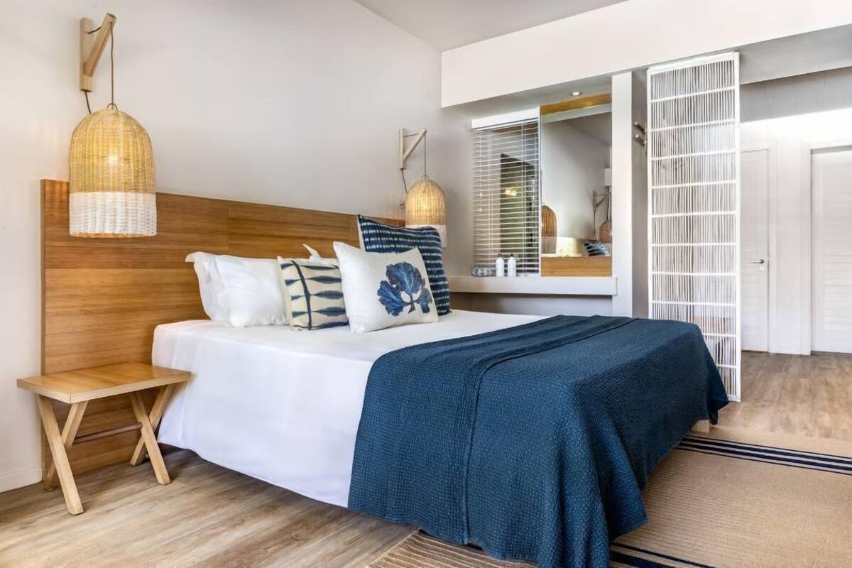 A modern bedroom with a wooden headboard, a bed with white bedding and a blue throw, a small wooden stool, and a large mirror.