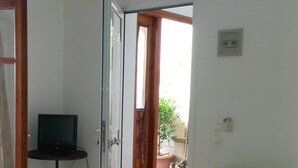 A room with a yellow sofa, a television on a small table, and a door leading to an outdoor area with plants. - Green Hill Studios & Apartments (Samos)