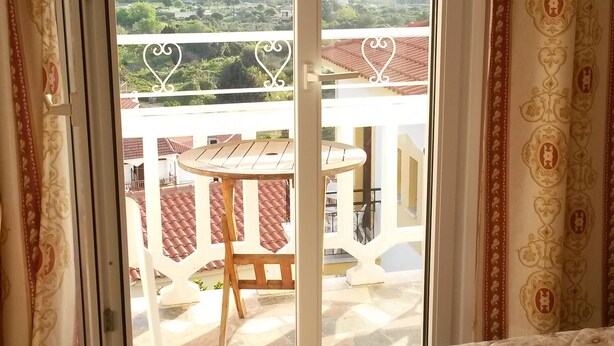 A bedroom with a bed, a balcony with a table and chairs, and a view of mountains. - Green Hill Studios & Apartments (Samos)