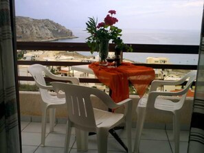 A balcony with a table and chairs, a vase with flowers, and a view of the sea and buildings.