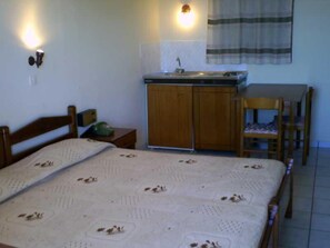 A bedroom with a bed, a wooden nightstand, a chair, a small table, a cabinet, and a window with curtains. - Yiannis (Karpathos)