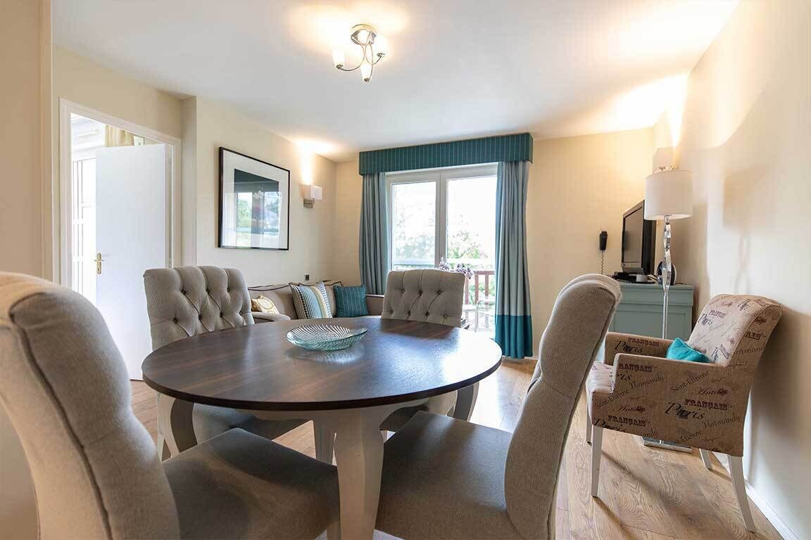A dining area with a round table, four upholstered chairs, a blue and white patterned armchair, a framed picture on the wall, and a window with curtains.