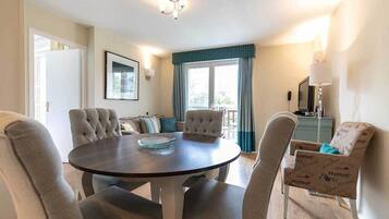 A dining area with a round table, four upholstered chairs, a blue and white patterned armchair, a framed picture on the wall, and a window with curtains.