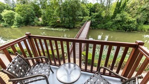 Une terrasse en bois avec deux chaises en métal et une table en verre, donnant sur une rivière traversée par un pont en bois.