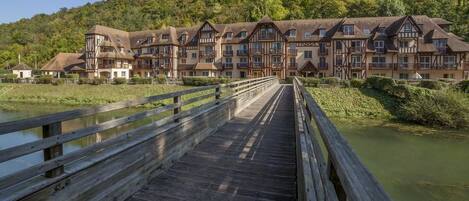 Un puente de madera que lleva a un hotel de estilo europeo tradicional, con varios balcones y un río al frente.