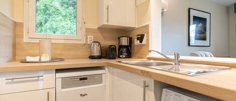 A modern kitchen with white cabinetry, a dishwasher, a coffee maker, and a window with a view of trees.