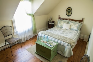 Une chambre à coucher avec un lit en bois, une table de chevet verte, un fauteuil en osier et une fenêtre avec des rideaux.
