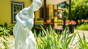 A white statue in a garden with a yellow building in the background. - Les Matins de Victoria (Waterloo)