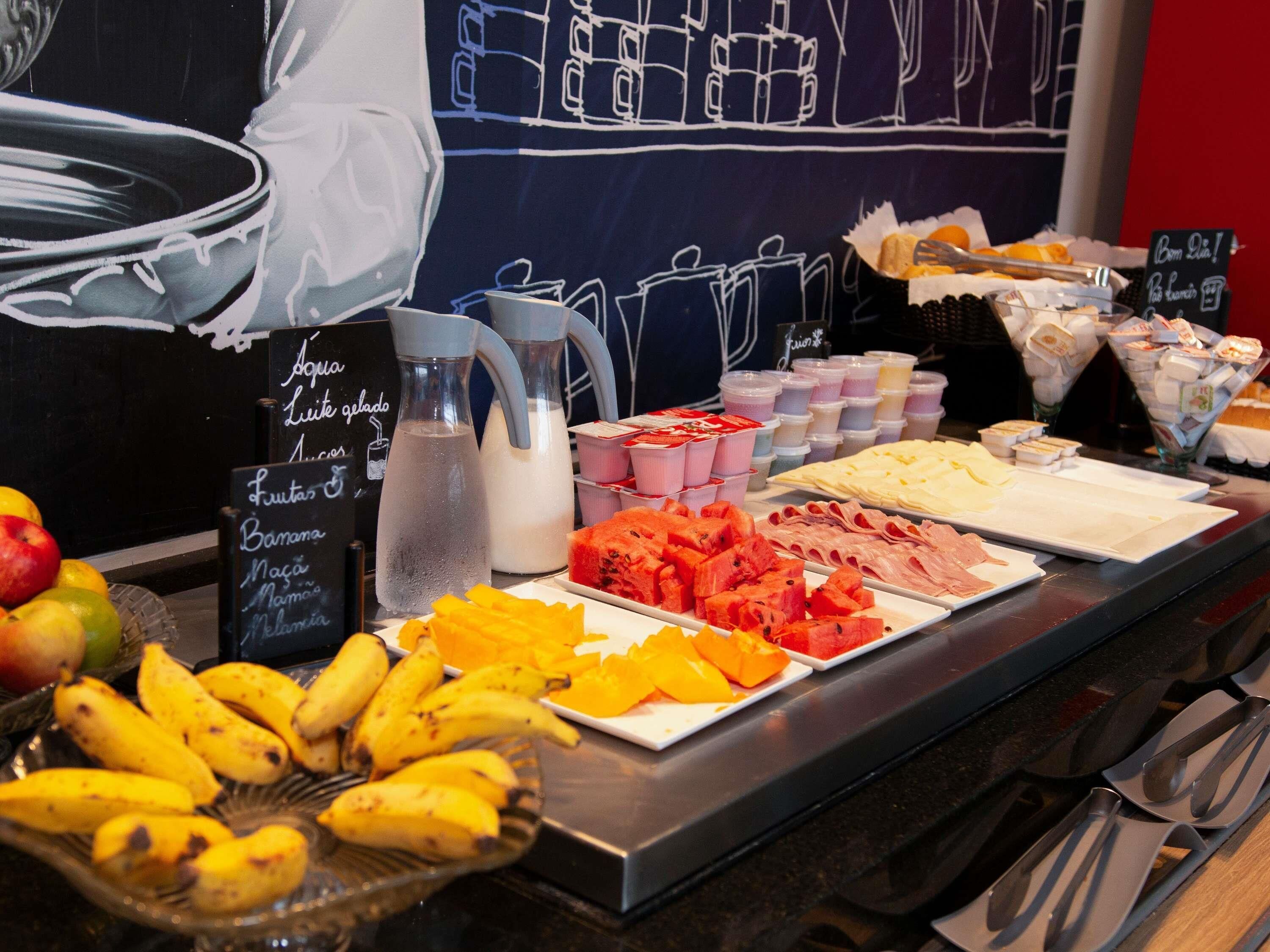 Un buffet proposant divers fruits, une carafe d’eau et un menu écrit à la craie sur un tableau noir.