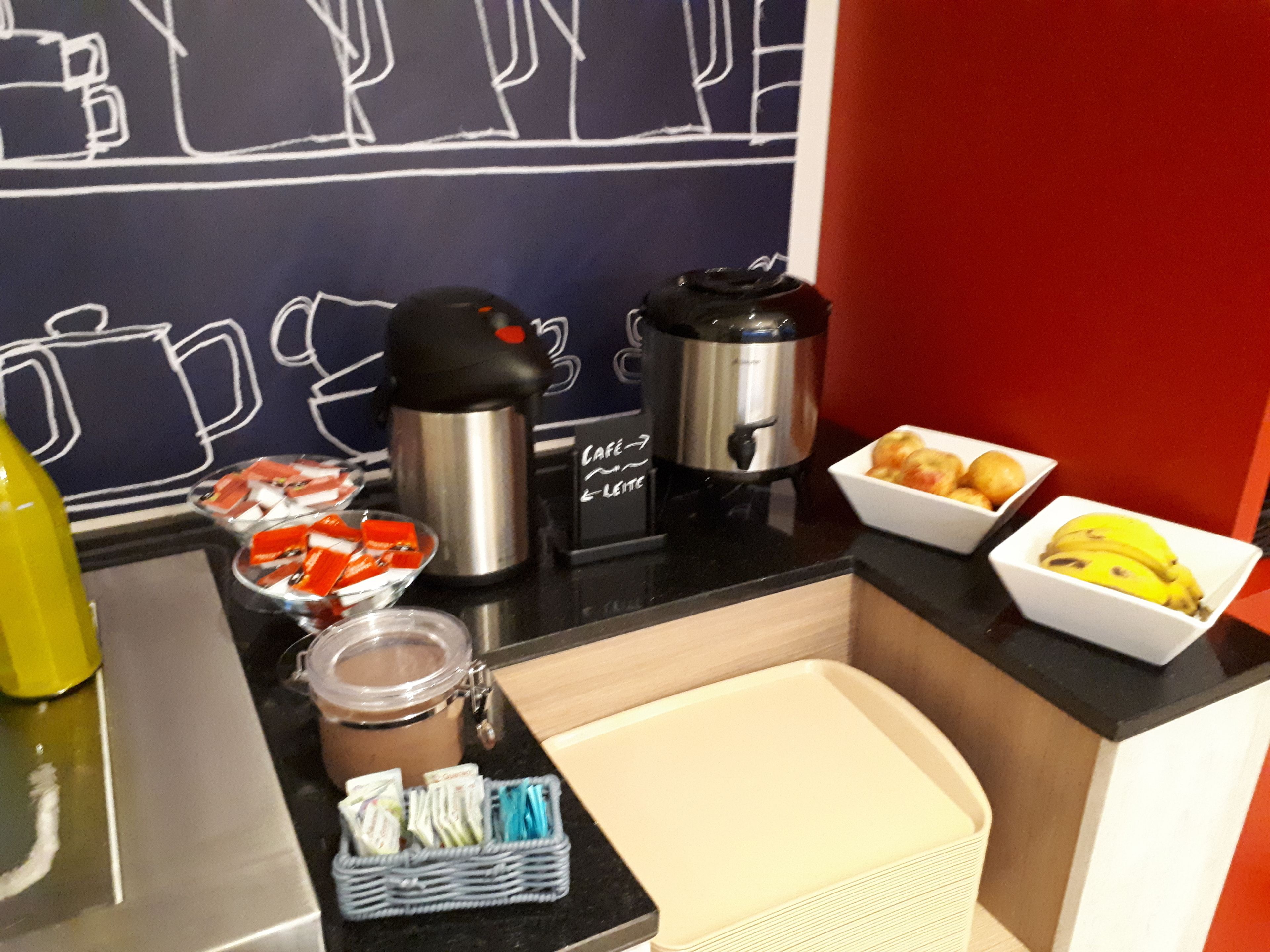 A breakfast bar with a coffee maker, a thermos, a jar of jam, a basket of pastries, and a bowl of fruit.