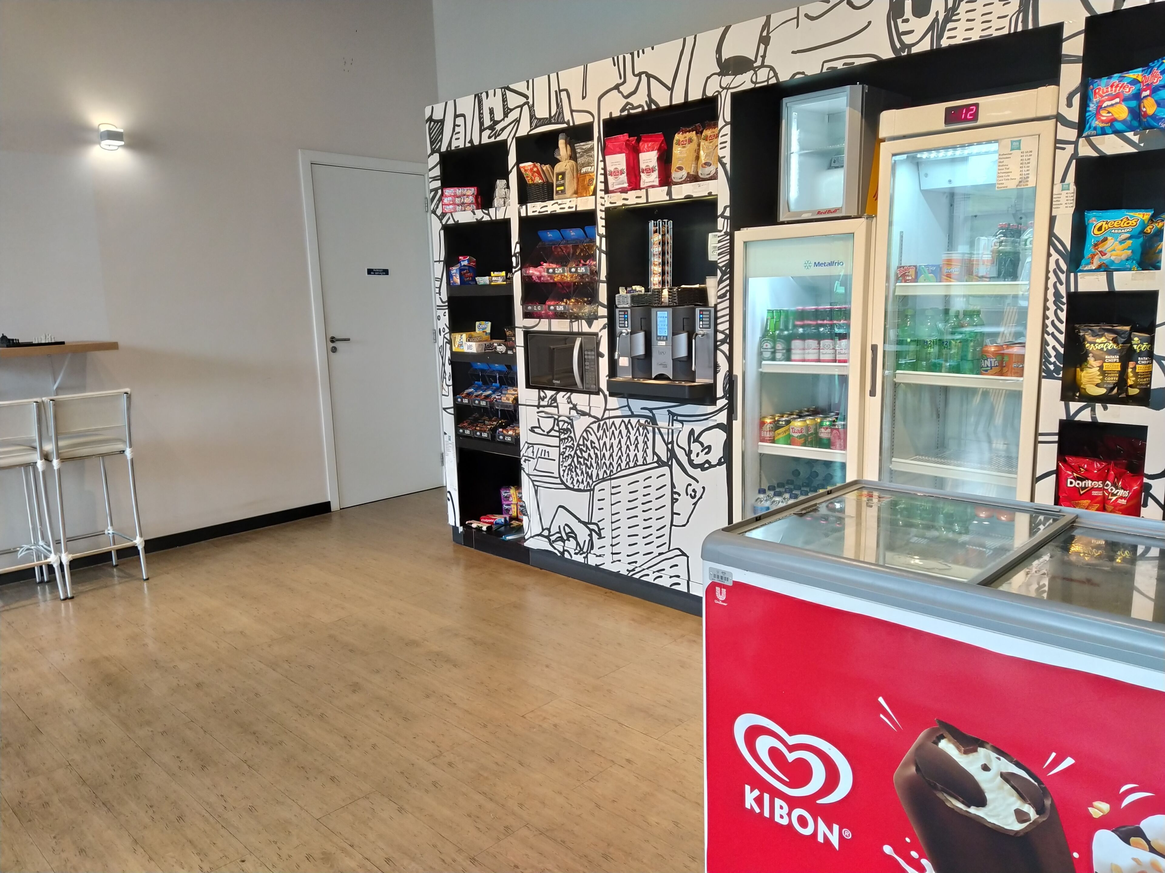 Espaço com máquinas de venda automática, diversas opções de lanches e bebidas, um refrigerador e uma vitrine de sorvetes Kibon.