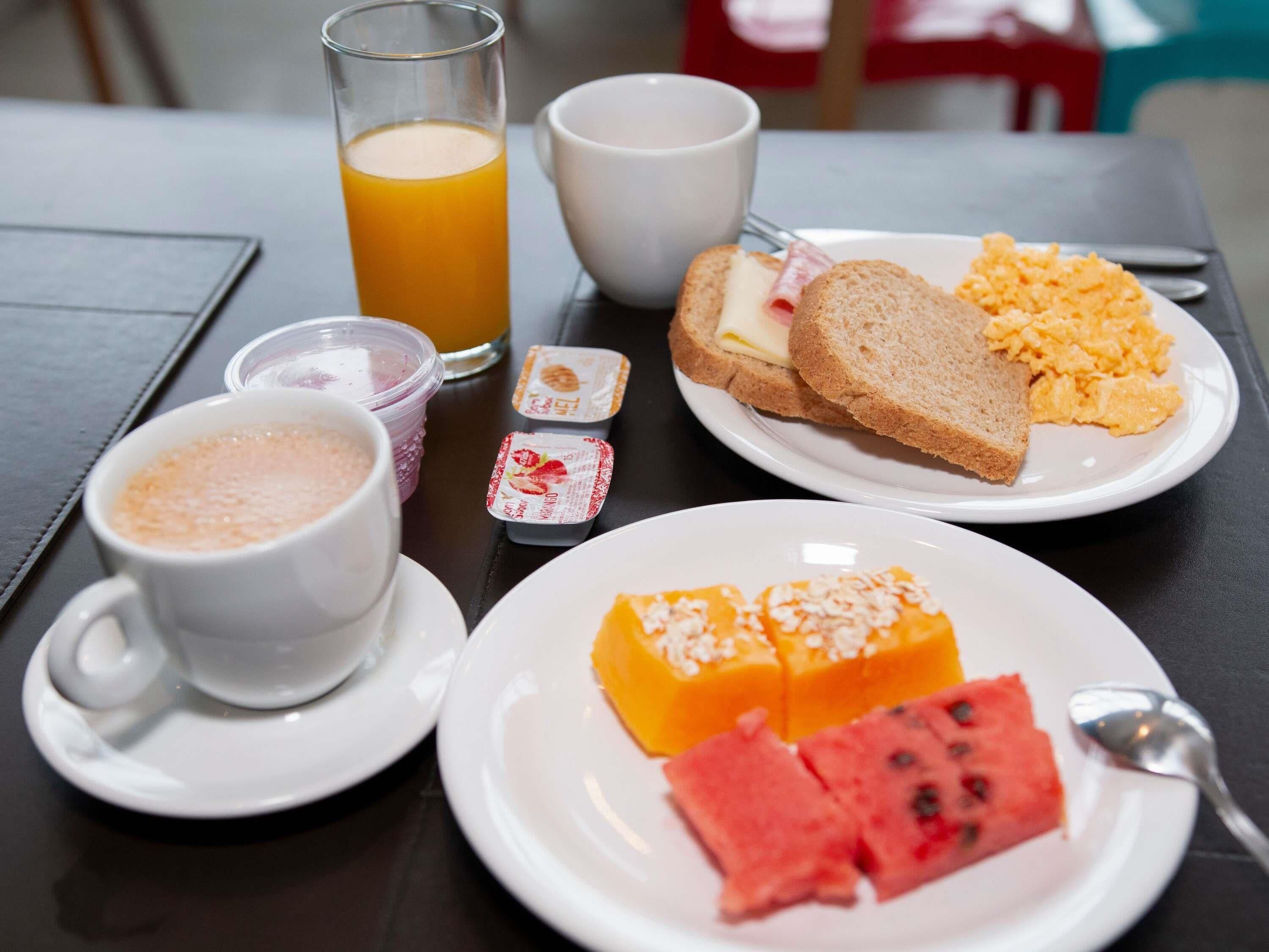 Uma mesa de café da manhã com torradas, ovos mexidos, um copo de suco de laranja, uma xícara de café, e um prato com melancia e queijo.