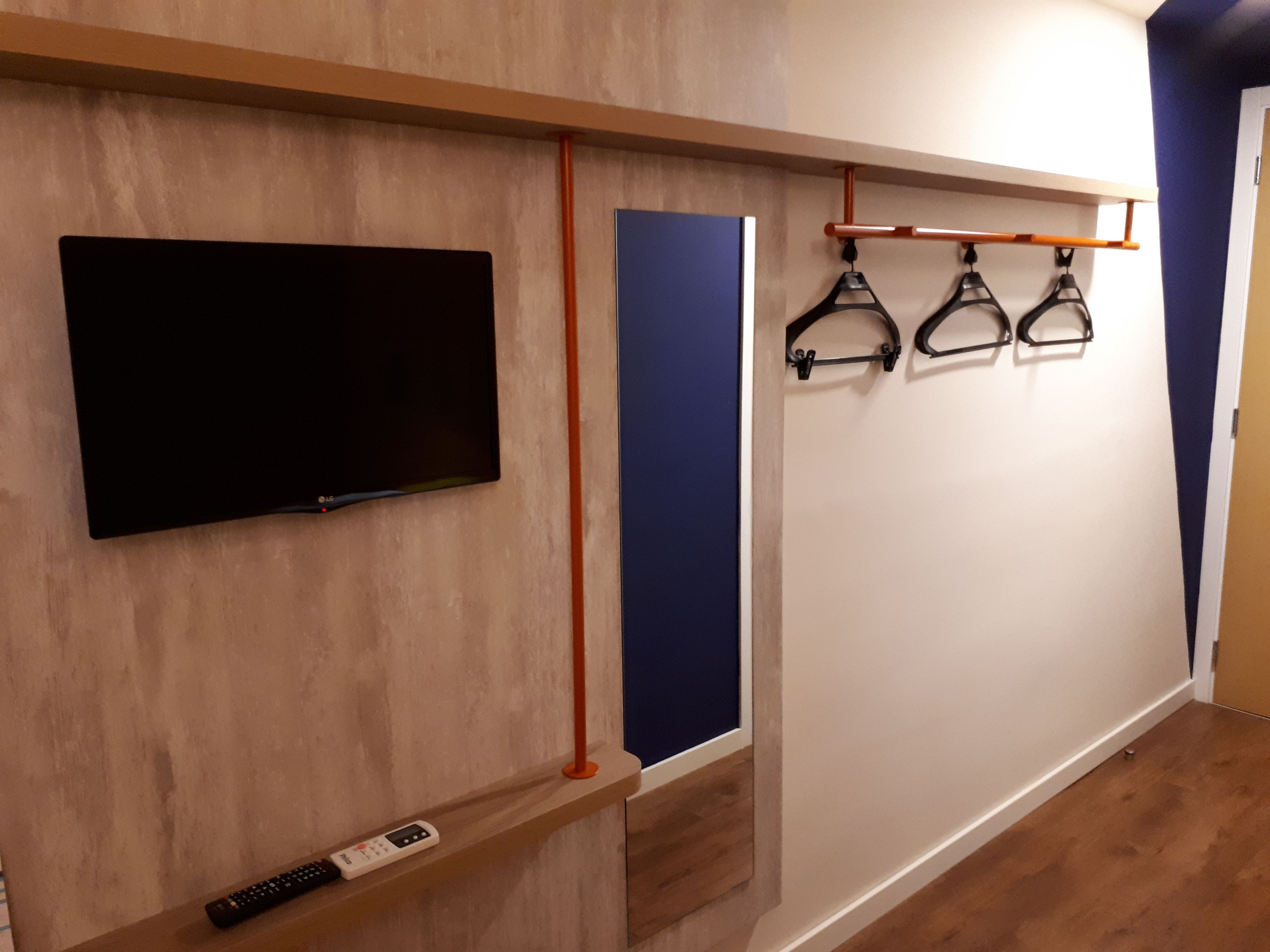 Une pièce avec un téléviseur fixé au mur, une étagère avec une télécommande et un porte-objets mural avec des articles suspendus.
