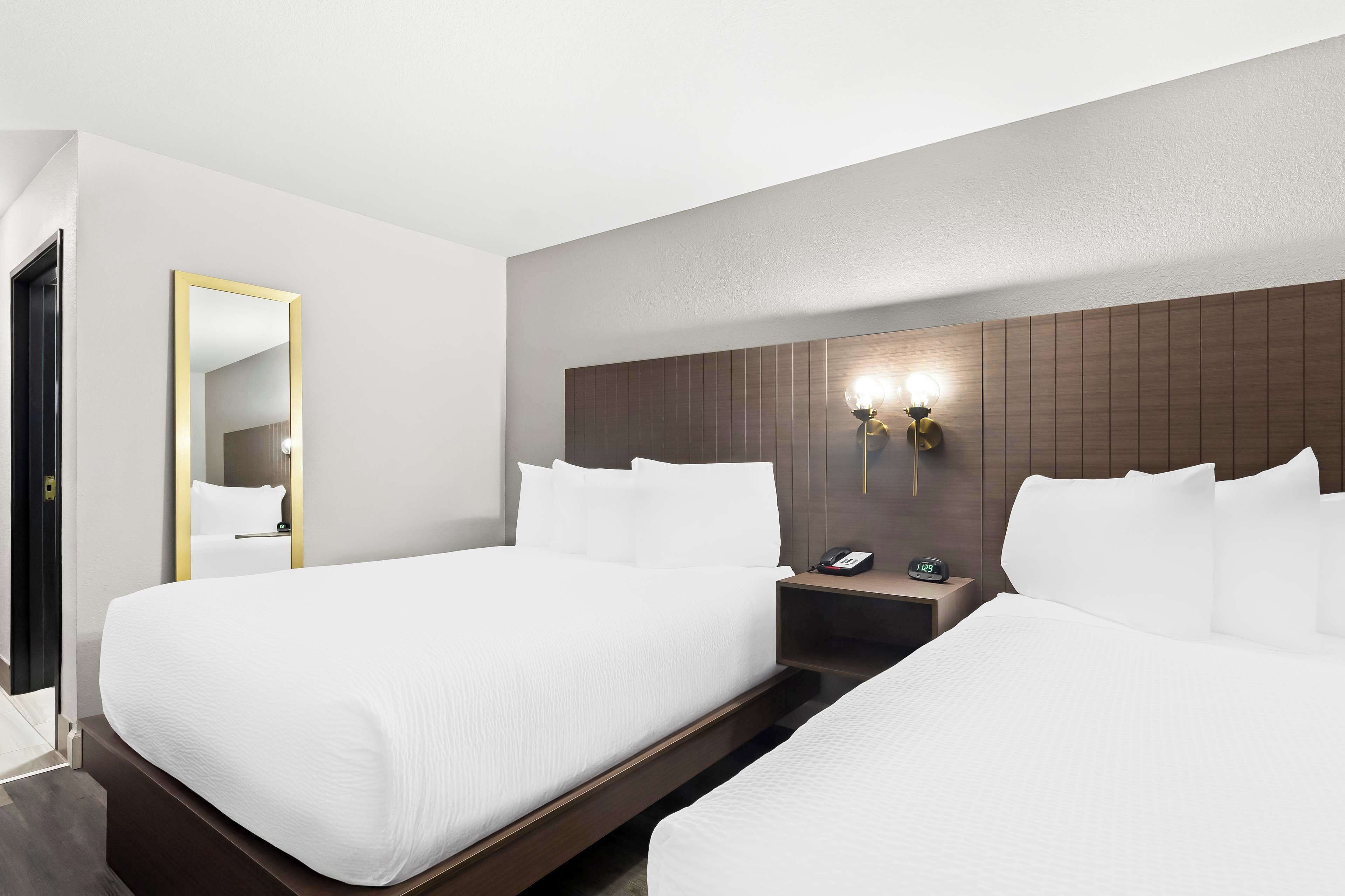 Une chambre d’hôtel avec deux lits, une table de chevet avec un téléphone et un réveil, un miroir et un luminaire fixé au mur.