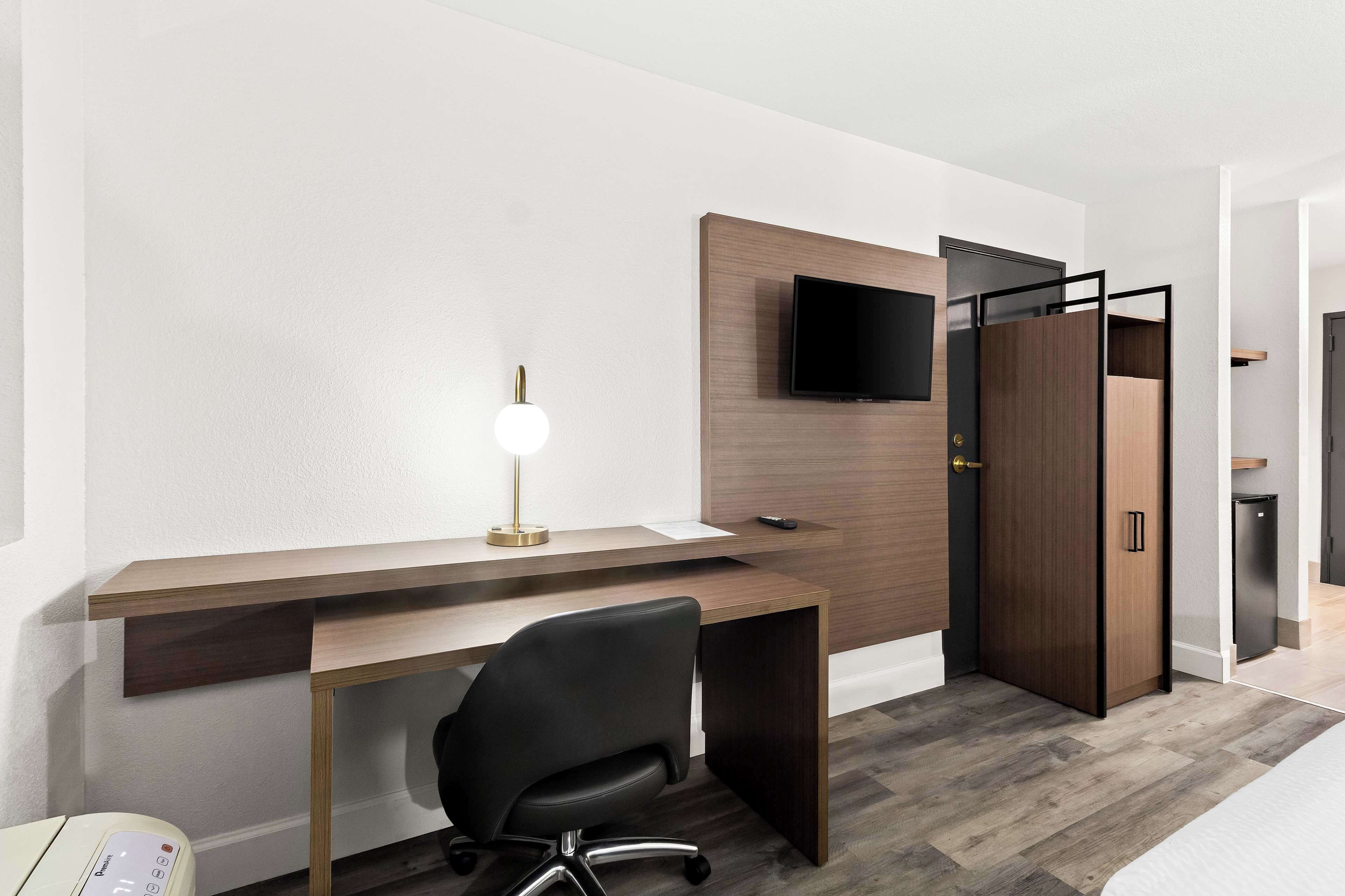 Una habitación de hotel moderna con un escritorio de madera, una silla de oficina negra, un televisor de pantalla plana montado en la pared y un armario con puerta corredera.