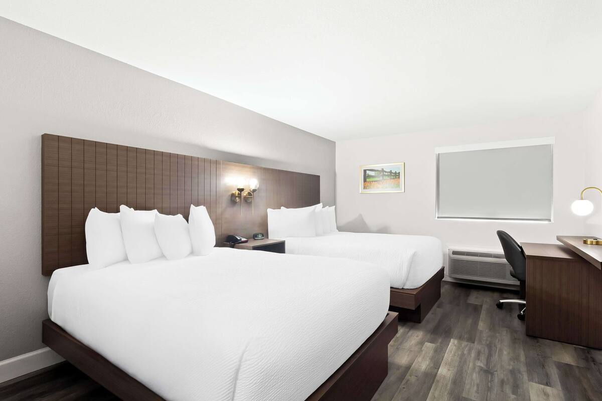 A hotel room with two beds, a desk, a chair, a nightstand, a wall-mounted lamp, and a picture on the wall.