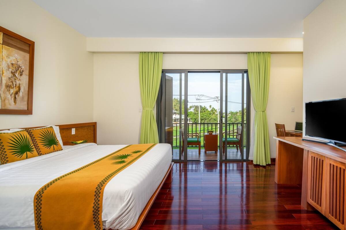 A hotel room with a bed, a TV, a desk, and a balcony with a view of greenery.