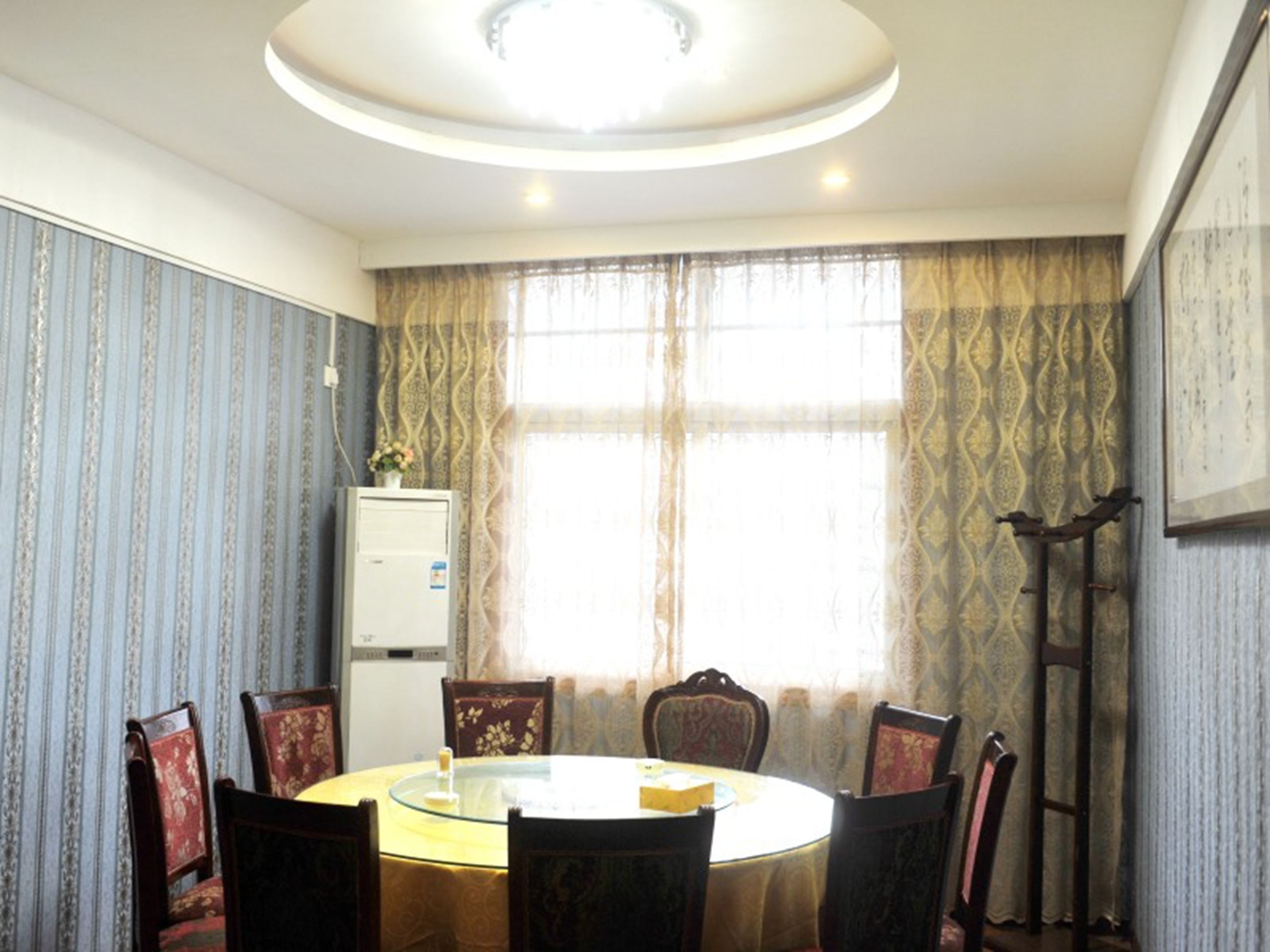 A conference room with a round table, six chairs, a white air conditioner, patterned curtains, and a ceiling light fixture.