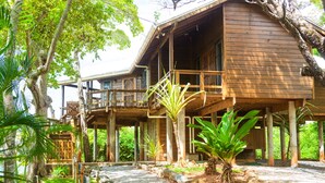 Una casa de fusta amb un balcó, envoltada de vegetació tropical i un camí de grava.