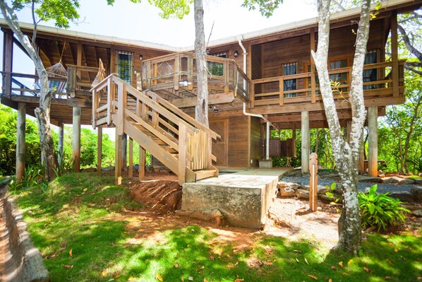 Une cabane en bois perchée dans un arbre, accessible par un escalier menant à une terrasse, le tout entouré d’une végétation luxuriante.