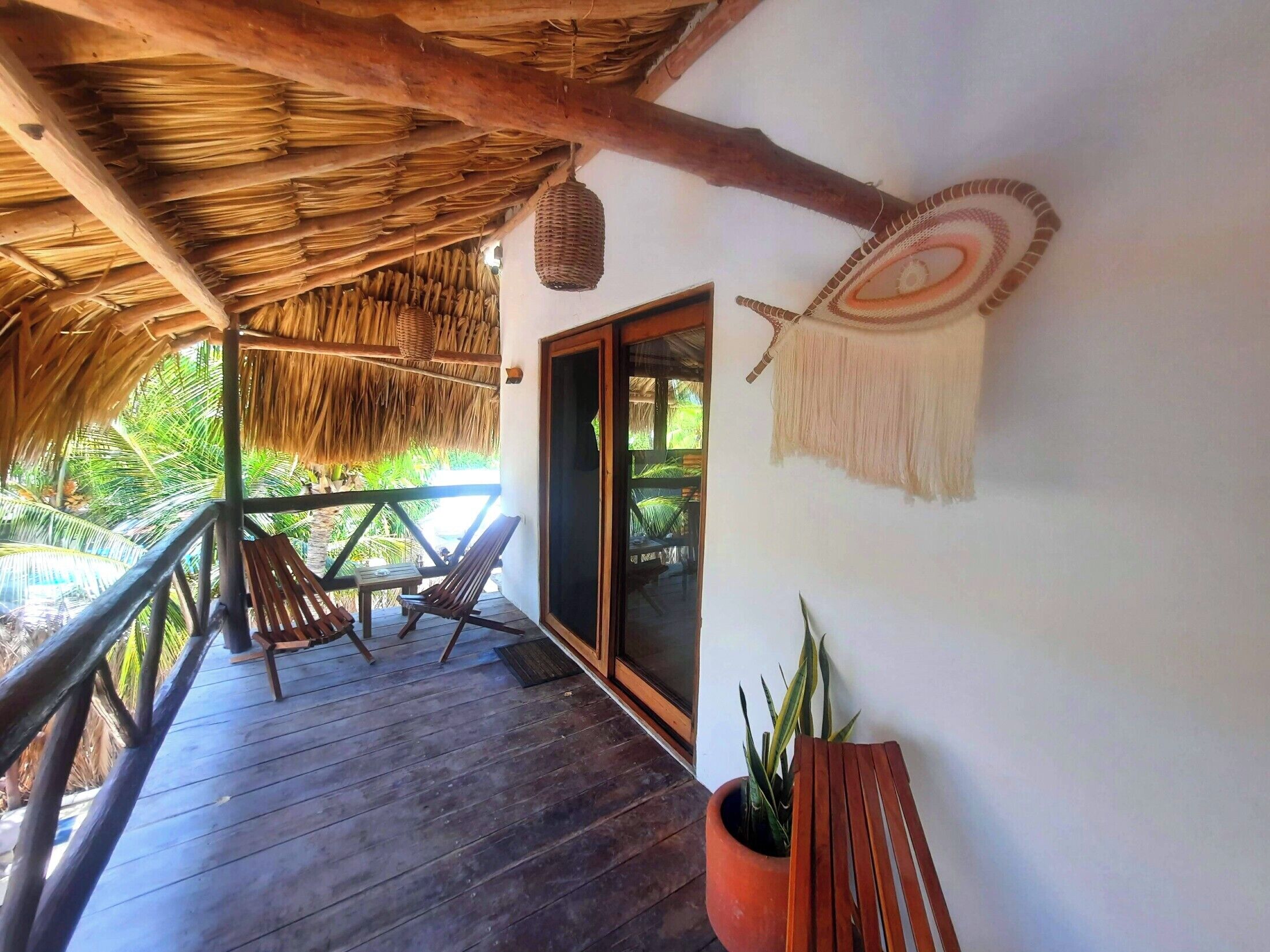 Una terraza de madera con techo de paja, un banco de madera, una planta en maceta y un adorno fijado a la pared.