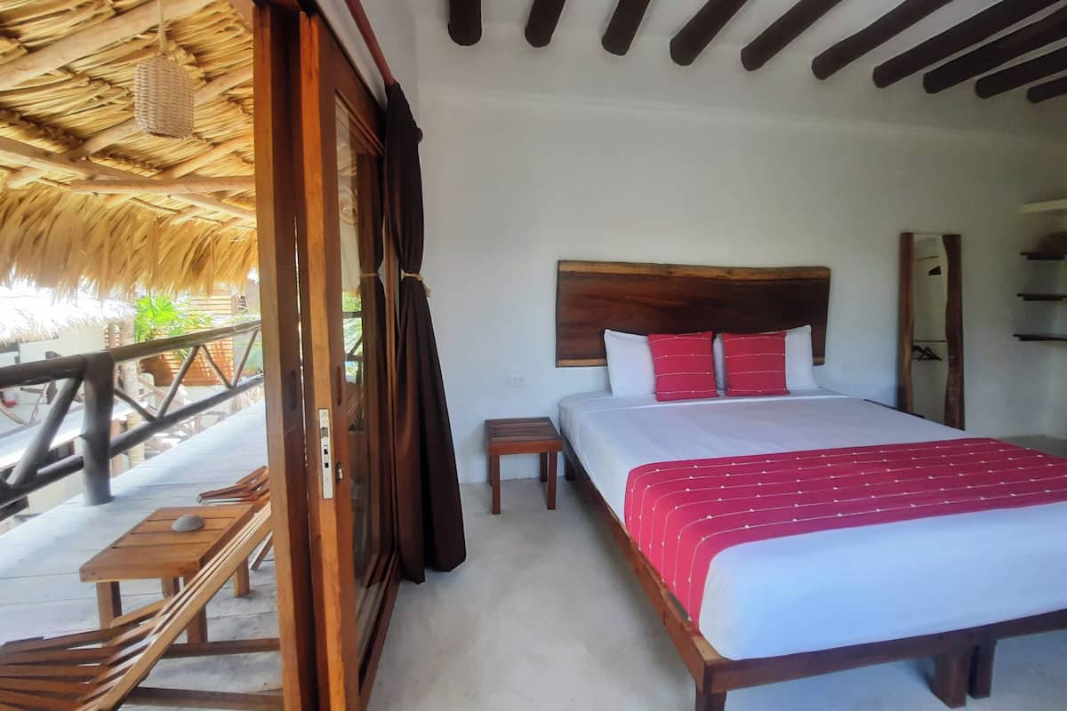 Un dormitorio con cabecera de madera, cama con ropa de cama blanca y roja, una mesita de noche pequeña y vistas al exterior a través de una puerta abierta.