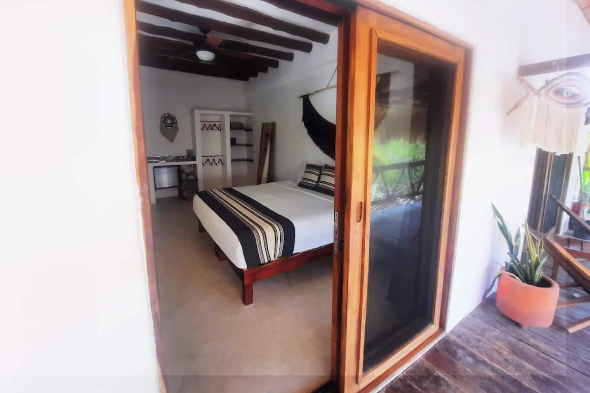 Un dormitorio con cama, escritorio, armario y puerta corrediza de vidrio que da a un espacio exterior con una planta en maceta.