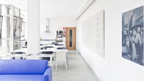 Une pièce moderne et bien éclairée, avec des murs blancs, un canapé bleu et un coin repas comprenant des tables et des chaises. De grandes œuvres d’art ornent les murs, et une porte donne accès à une autre pièce.
