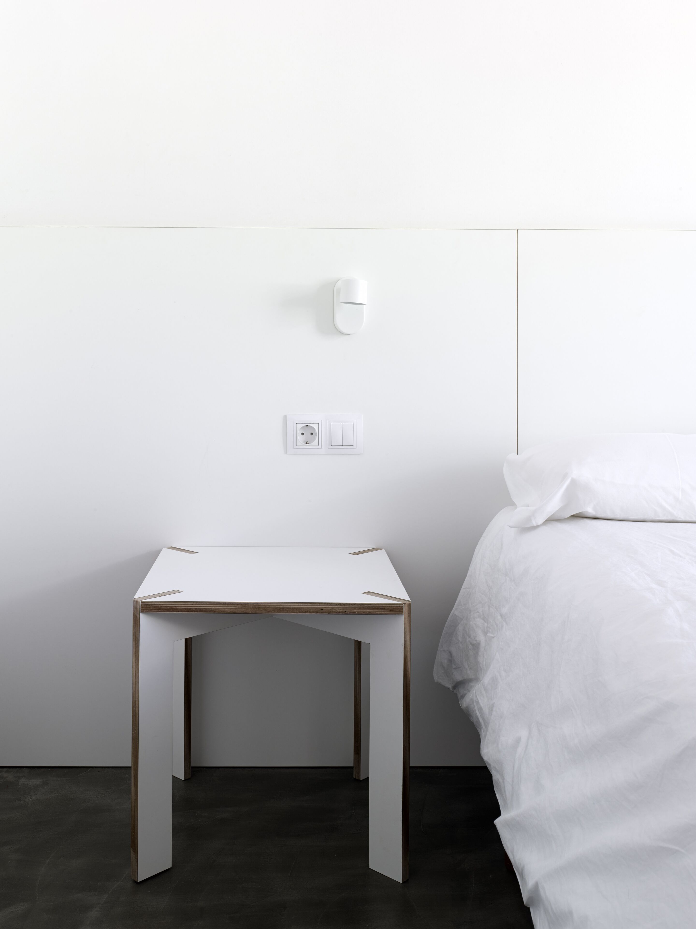Habitación minimalista con cama blanca, una mesita de noche pequeña también blanca y una lámpara de pared, además de un enchufe.